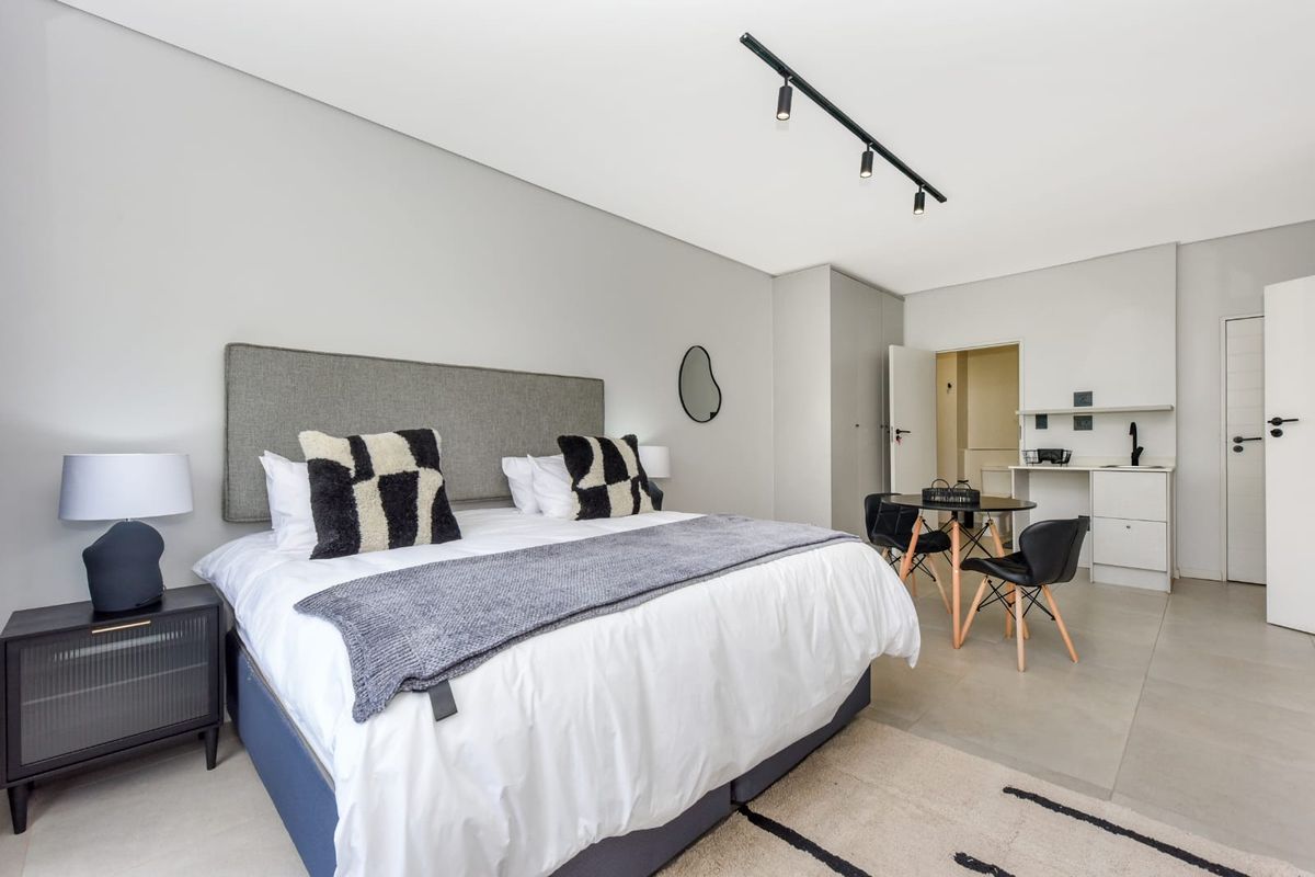 Modern bedroom featuring built-in cupboards, track lighting, and a small kitchenette area with tiled flooring throughout.