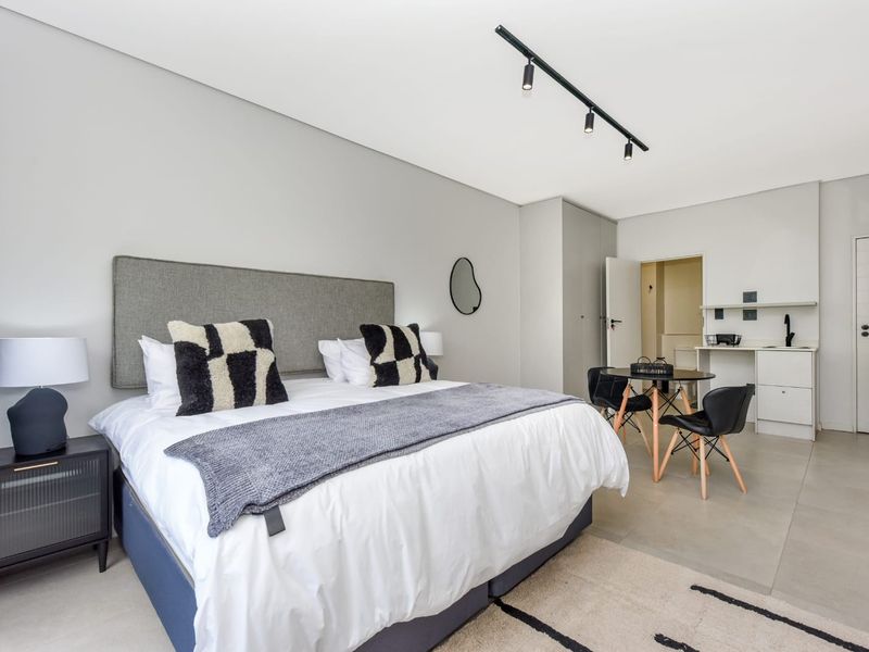 Modern bedroom featuring built-in cupboards, track lighting, and a small kitchenette area with tiled flooring throughout.
