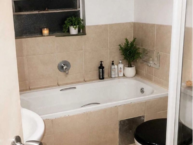 A compact bathroom featuring a built-in bathtub with wall-mounted mixer, tiled surround, and a standard toilet.