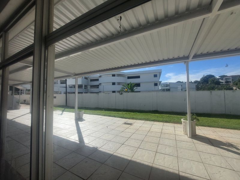 A covered outdoor patio area featuring tiled flooring, metal support columns, and a corrugated roof, overlooking a private garden space.