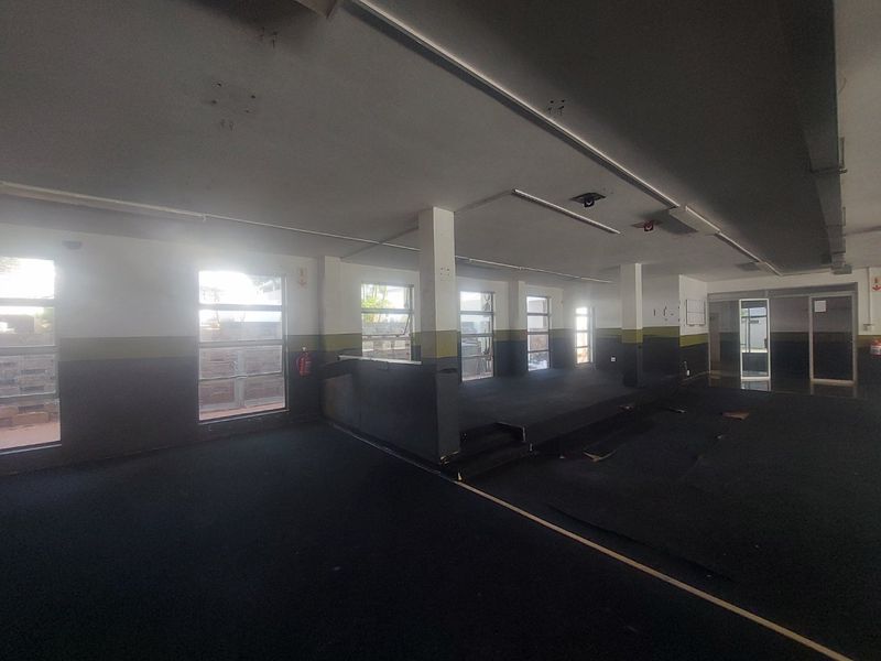 Spacious open-plan commercial area featuring a raised platform, large windows for natural light, and worn carpet flooring in need of refurbishment.
