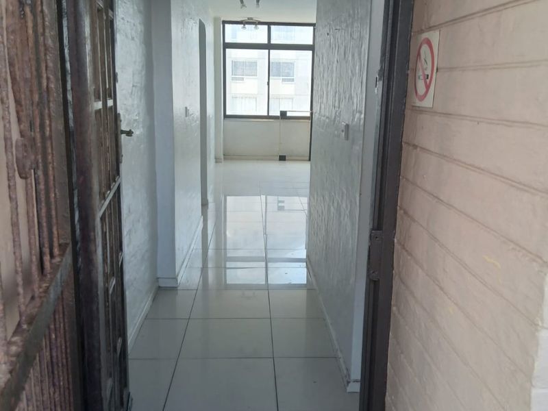A bright, empty hallway featuring tiled flooring and a metal security gate, leading towards a windowed living area.