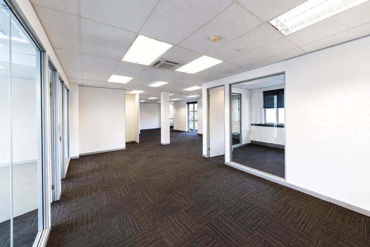 A spacious, vacant commercial office suite featuring modern glass partitioning, carpet tile flooring, and a suspended ceiling with integrated lighting and air conditioning.