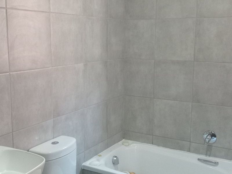 A clean, modern bathroom featuring neutral grey wall and floor tiles, a built-in bathtub, a white toilet, and a vanity with a basin.