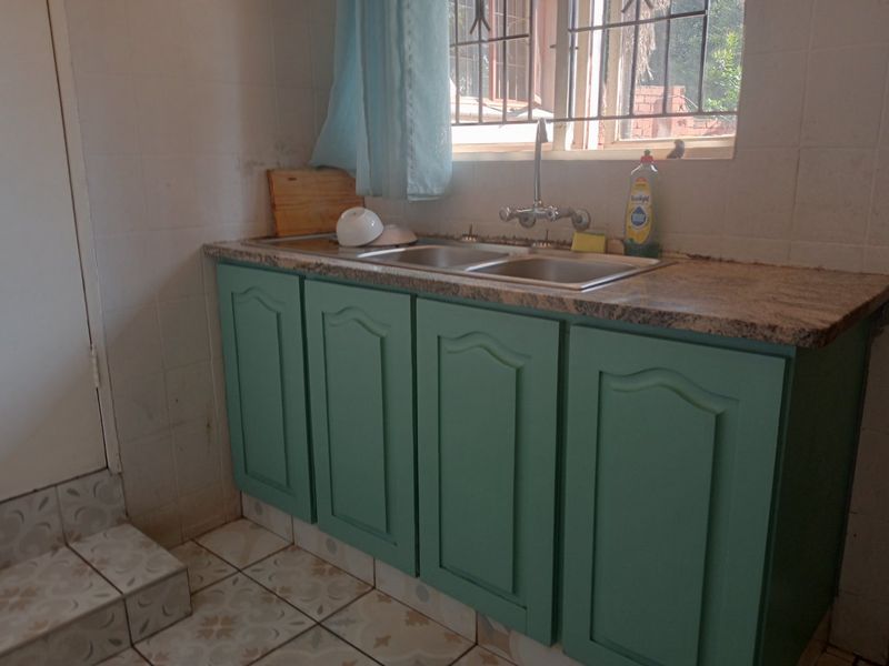 A functional kitchen area featuring a double basin stainless steel sink, granite countertops, and green cabinetry with tiled splashbacks and flooring.