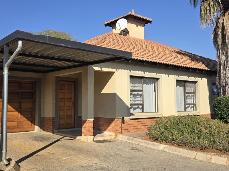 Front elevation of a townhouse featuring a tiled roof, a single carport, a wooden garage door, and a paved driveway with established garden beds.