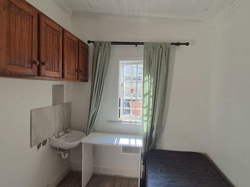 A compact bedroom featuring a wall-mounted basin, overhead wooden cabinetry, and security bars on the window, requiring cosmetic updates to improve the finish.