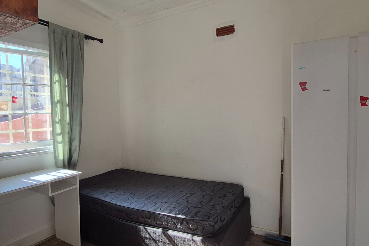 A compact bedroom featuring timber-look flooring, a multi-pane window, and a wall-mounted ventilation brick, offering a functional space with potential for cosmetic updates.