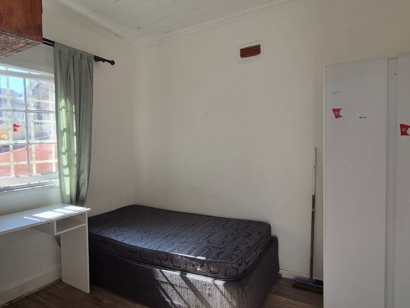 A compact bedroom featuring timber-look flooring, a multi-pane window, and a wall-mounted ventilation brick, offering a functional space with potential for cosmetic updates.