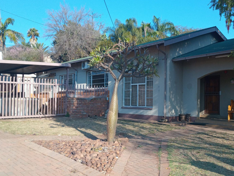 Front elevation of a single-story house featuring a paved driveway, covered carport, security fencing, and established garden area.