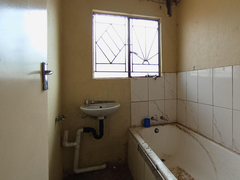 A dated bathroom featuring a wall-mounted basin, bathtub with tiled splashback, and security burglar bars on the window, requiring significant renovation and repair.