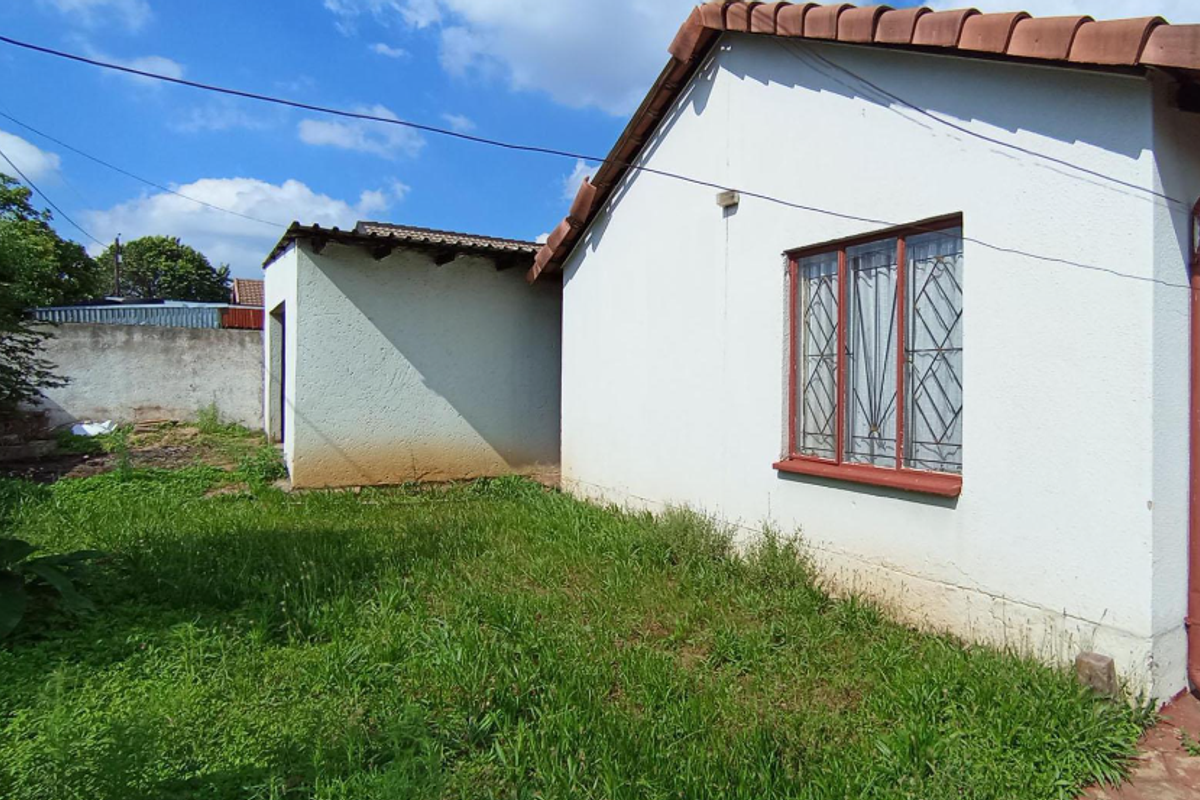 Rear view of the property showing the exterior wall, security-barred window, and a small backyard area with overgrown grass.