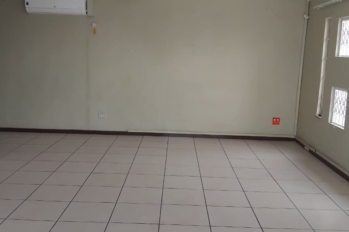 An empty open-plan living area featuring tiled flooring, a wall-mounted air conditioning unit, and windows fitted with security bars.