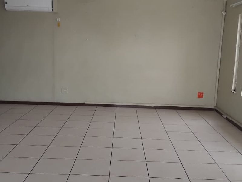 An empty open-plan living area featuring tiled flooring, a wall-mounted air conditioning unit, and windows fitted with security bars.