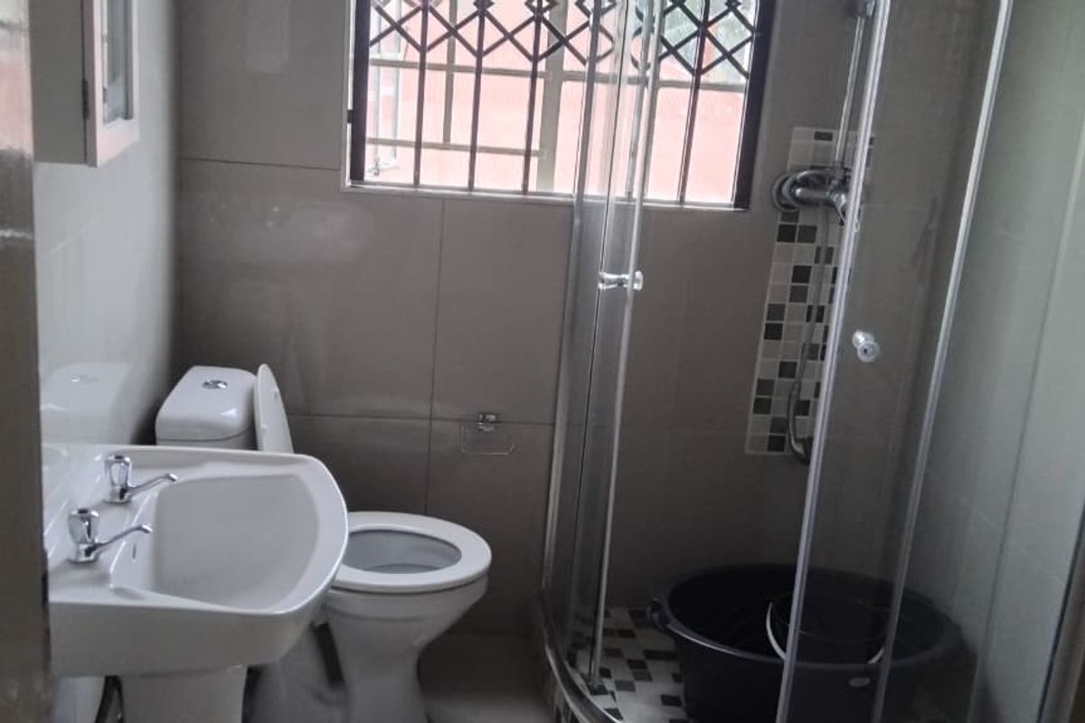 A functional bathroom featuring a pedestal basin, toilet, and a corner shower enclosure with tiled walls and security bars on the window.
