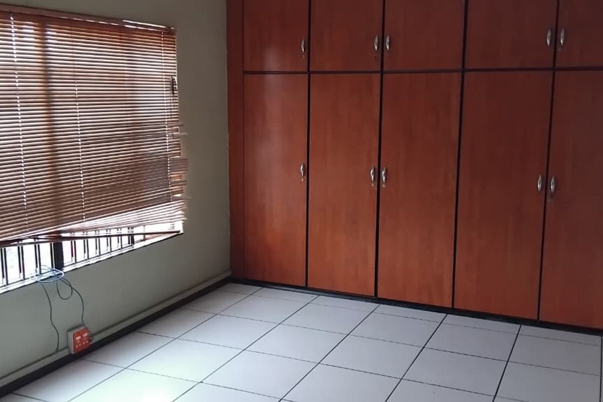 A vacant bedroom featuring tiled flooring, extensive built-in wooden cupboards, and a window fitted with venetian blinds and security bars.