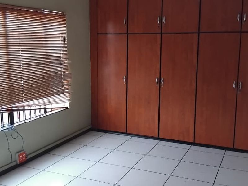 A vacant bedroom featuring tiled flooring, extensive built-in wooden cupboards, and a window fitted with venetian blinds and security bars.