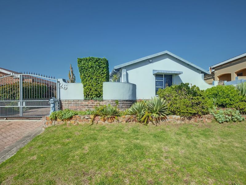 Front elevation of a single-story house featuring a paved driveway, metal security gate, and established garden with a brick boundary wall.
