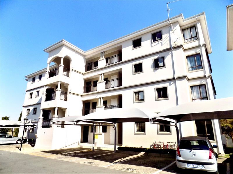 Exterior view of a multi-story apartment building featuring covered parking bays, private balconies, and a paved driveway area.