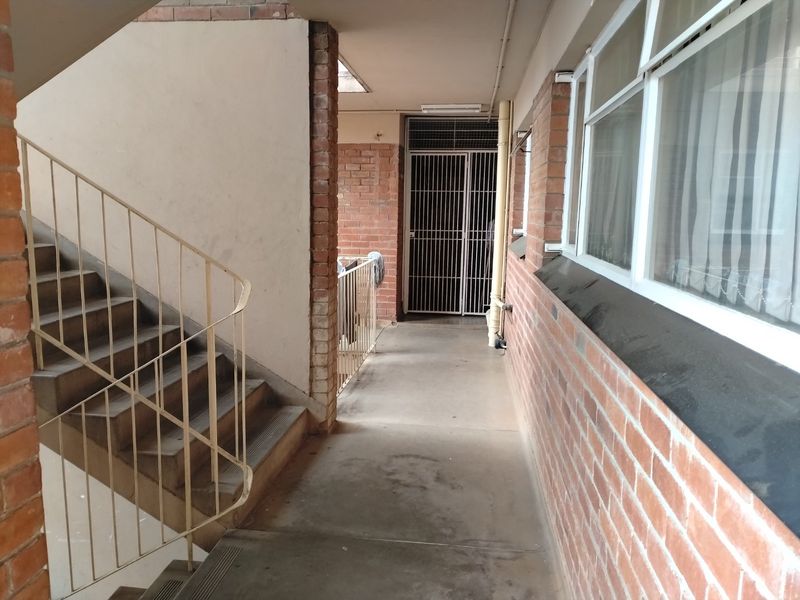An exterior apartment hallway featuring exposed brick walls, a concrete walkway, a metal staircase with railing, and a secure metal gate entrance.