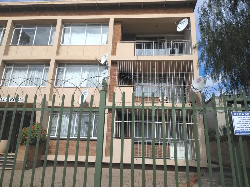 Front elevation of a multi-story apartment building featuring brick facade, private balconies with security bars, and perimeter fencing with razor wire.
