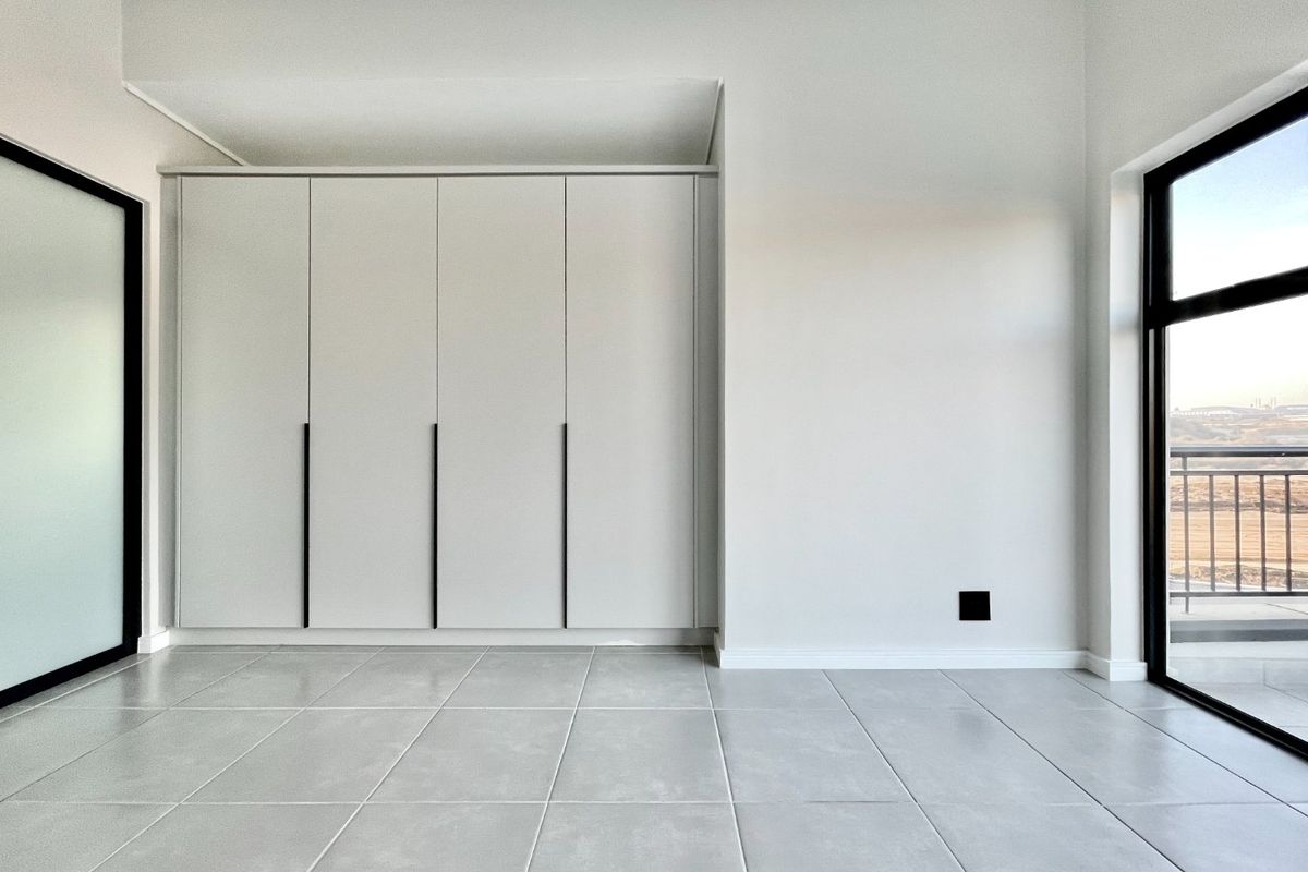 A bright, modern bedroom featuring neutral tiled flooring, floor-to-ceiling built-in cupboards, and a large sliding glass door leading to a balcony.