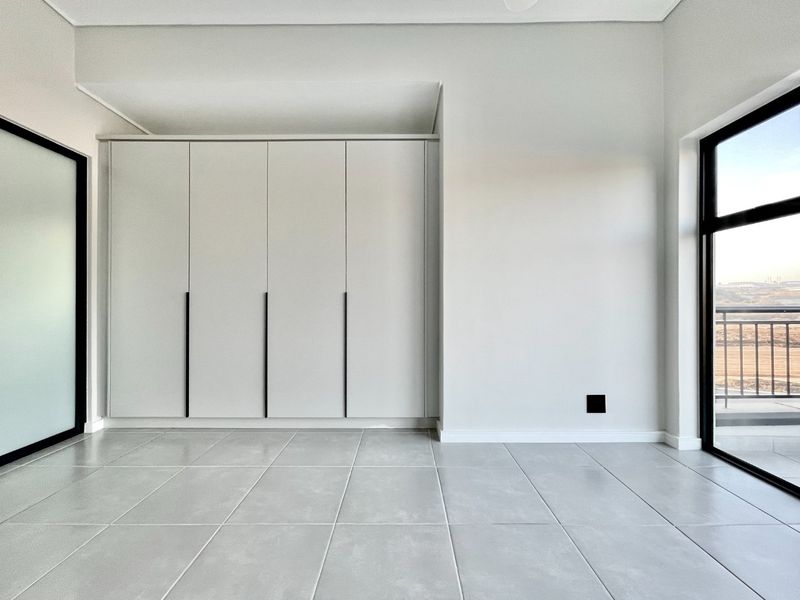 A bright, modern bedroom featuring neutral tiled flooring, floor-to-ceiling built-in cupboards, and a large sliding glass door leading to a balcony.
