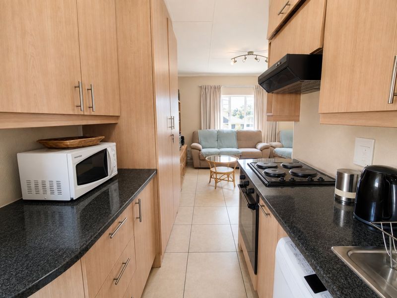 An open-plan kitchen and living area featuring light wood cabinetry, granite countertops, integrated appliances, and neutral tiled flooring.