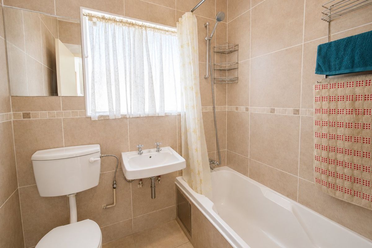 A functional bathroom featuring neutral ceramic wall and floor tiles, a wall-mounted basin, a close-coupled toilet, and a bathtub with a shower mixer.