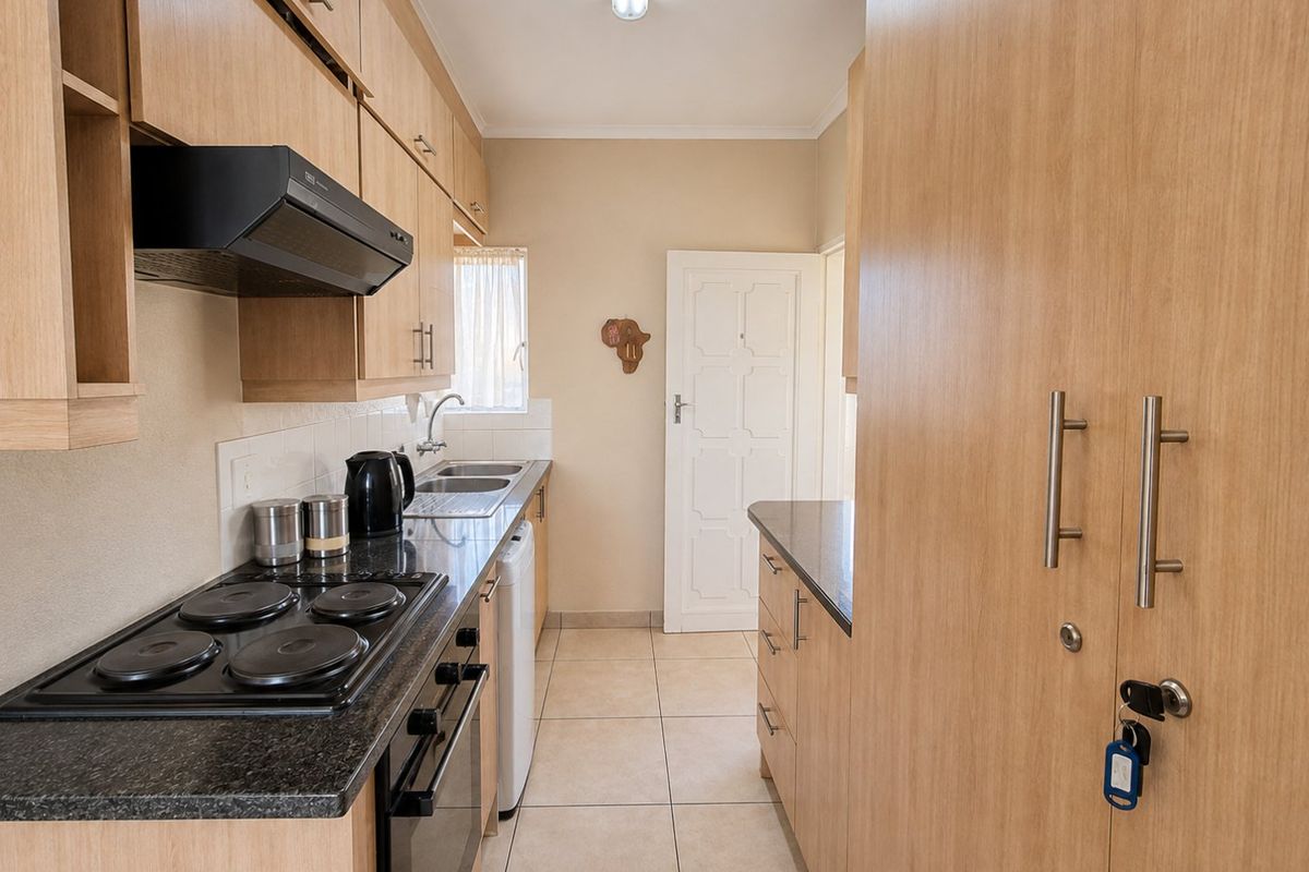 A functional galley-style kitchen featuring light wood cabinetry, dark laminate countertops, an integrated electric hob, oven, and extractor fan with tiled flooring.