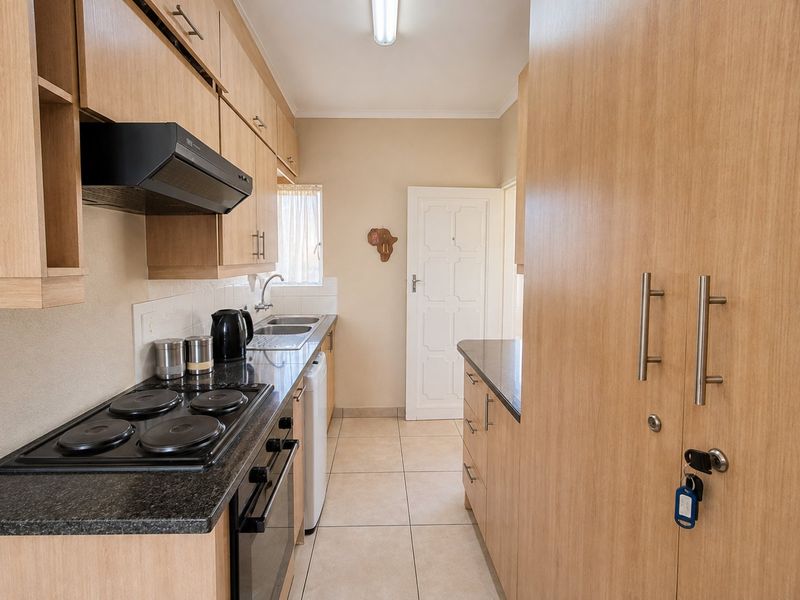 A functional galley-style kitchen featuring light wood cabinetry, dark laminate countertops, an integrated electric hob, oven, and extractor fan with tiled flooring.