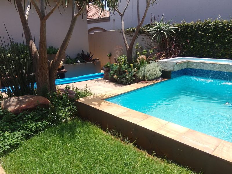 A well-maintained outdoor pool area featuring a tiled swimming pool with a built-in water feature, surrounded by paved patio and landscaped garden beds.