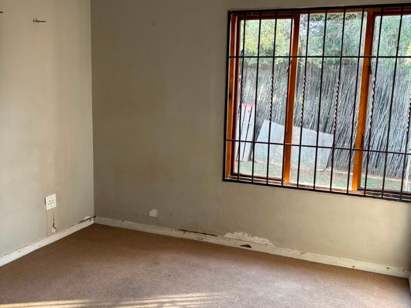 An empty bedroom featuring a ceiling fan, carpeted flooring, and security bars on the window. The room requires cosmetic updates to the walls and skirting.