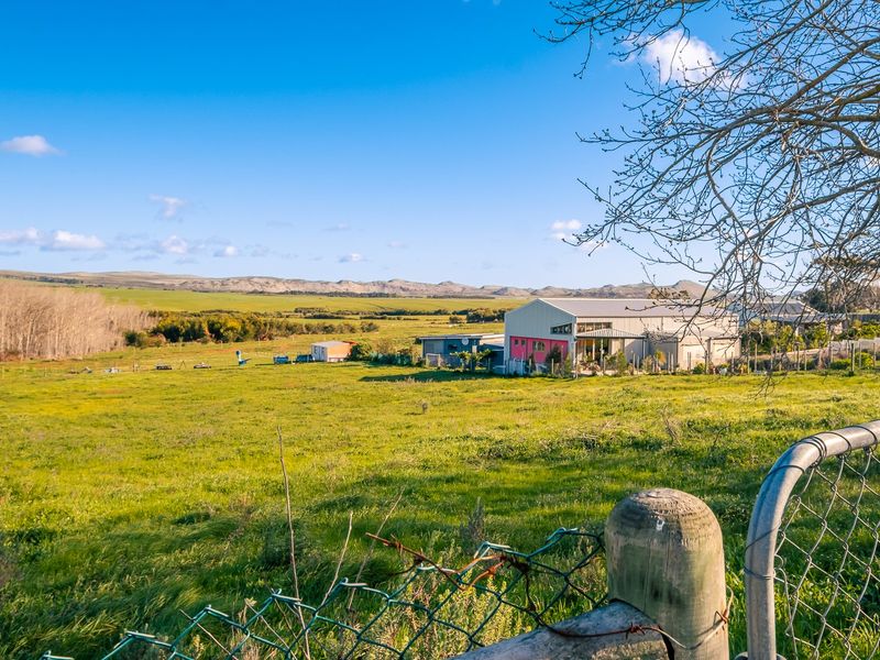 A wide-angle view of a large residential vacant land plot featuring open grassy fields, wire fencing, and distant rural buildings under a clear sky.