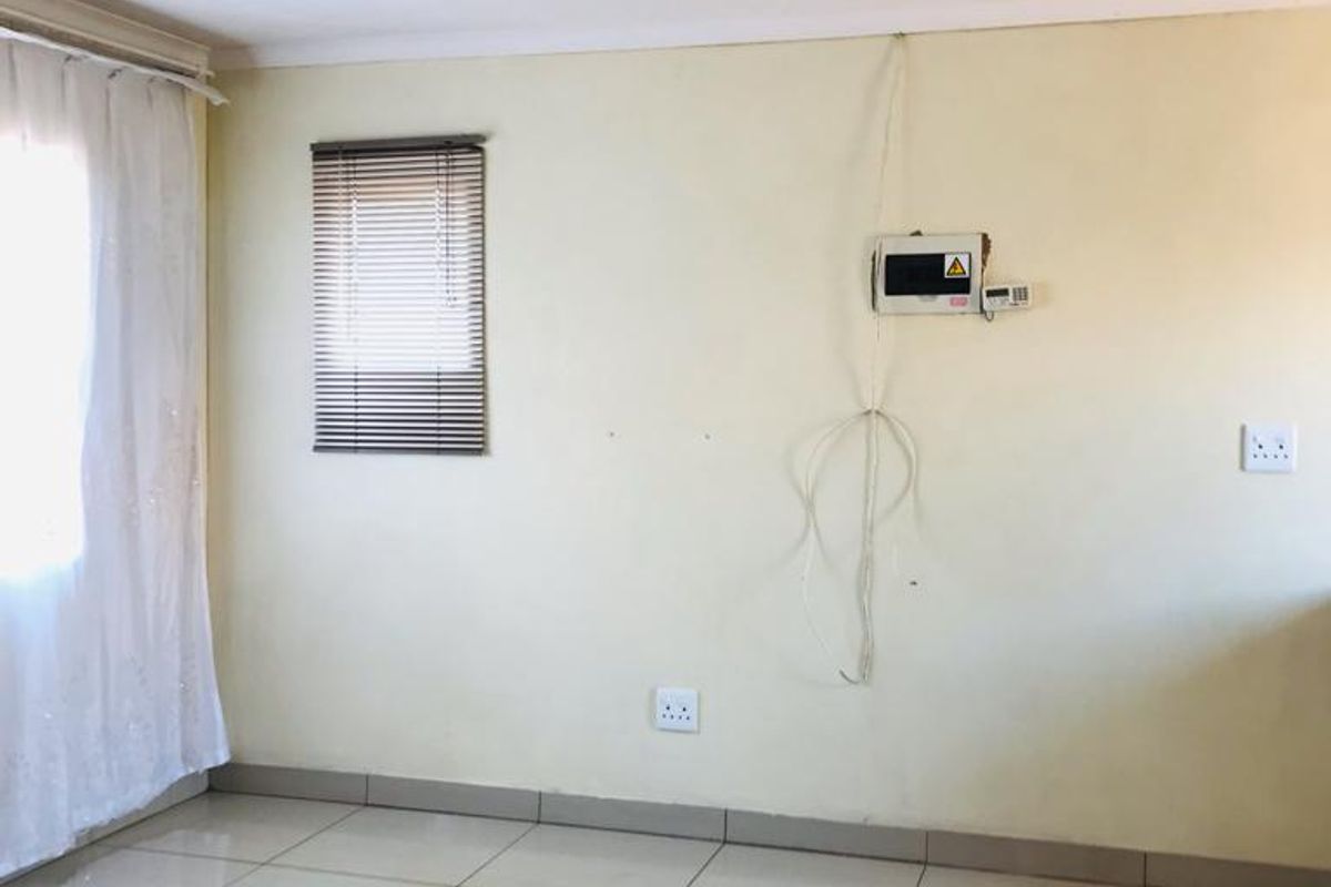 An empty living area featuring tiled flooring, venetian blinds, and wall-mounted security infrastructure. The space requires minor cosmetic updates and cable management.