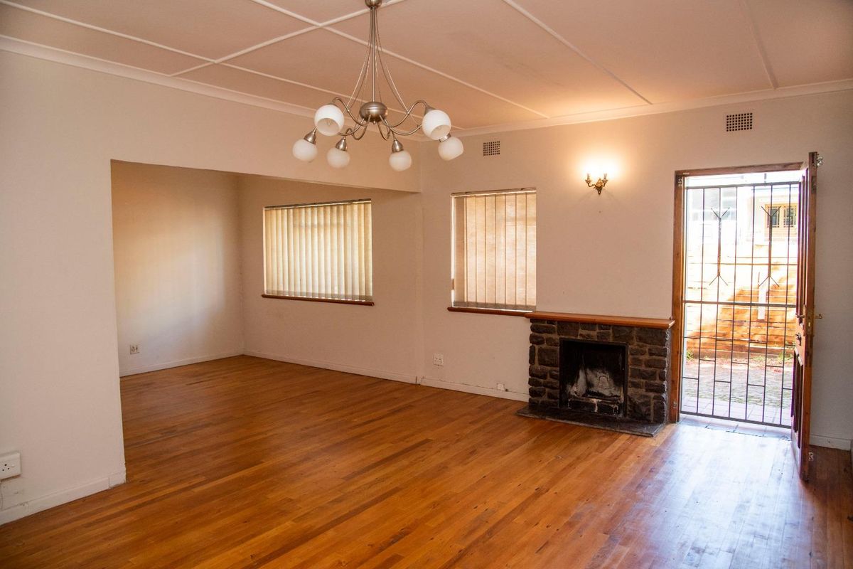 Spacious living room featuring original hardwood flooring, a classic stone fireplace, and natural light through vertical blinds, offering a versatile open-plan layout.