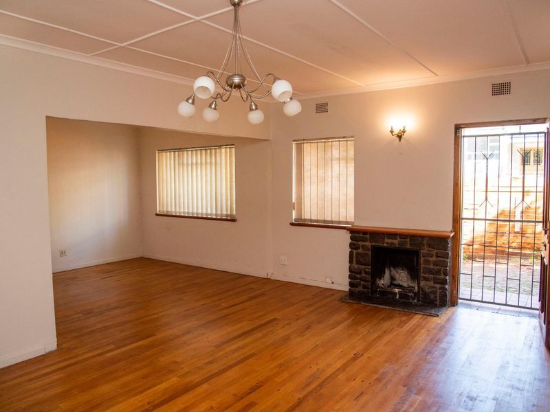 Spacious living room featuring original hardwood flooring, a classic stone fireplace, and natural light through vertical blinds, offering a versatile open-plan layout.