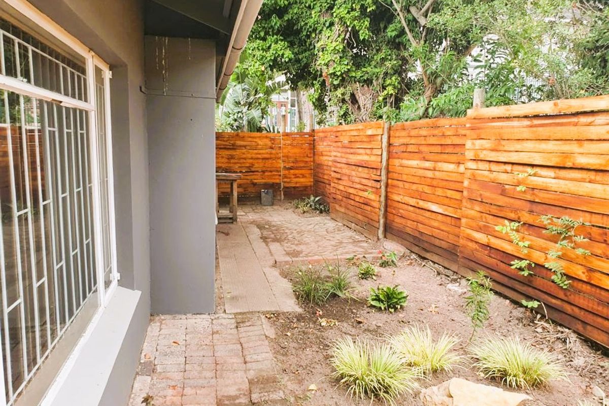 A private rear garden area featuring timber slat fencing, brick paving, and established garden beds alongside the side of the house.