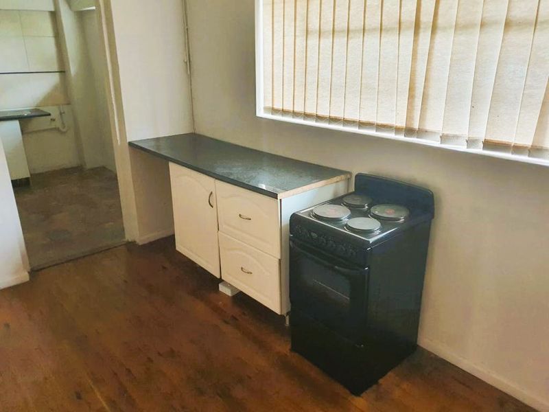 A dated kitchen area featuring hardwood flooring, base cabinetry with laminate countertops, and a freestanding electric stove, with an adjacent scullery or pantry space.