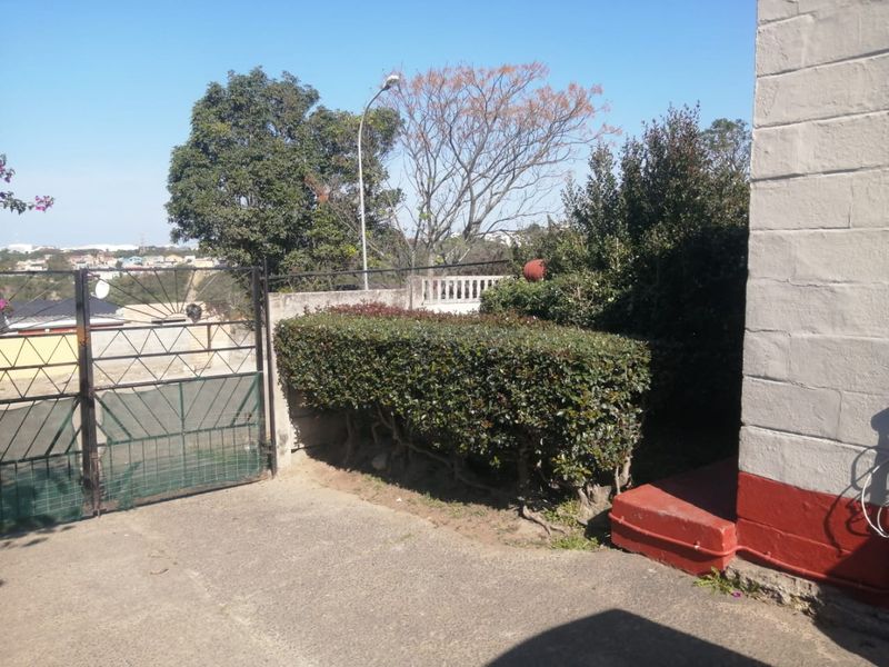 Front exterior view showing a metal driveway gate, concrete driveway, and established hedging alongside the property boundary wall.