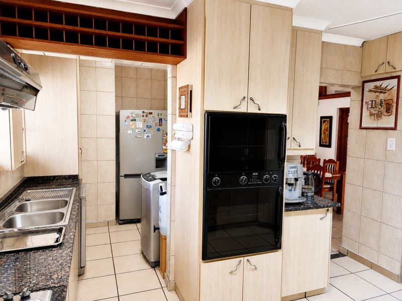 A functional kitchen featuring light wood cabinetry, granite countertops, a built-in oven and microwave, and tiled finishes throughout, with an adjacent scullery area.