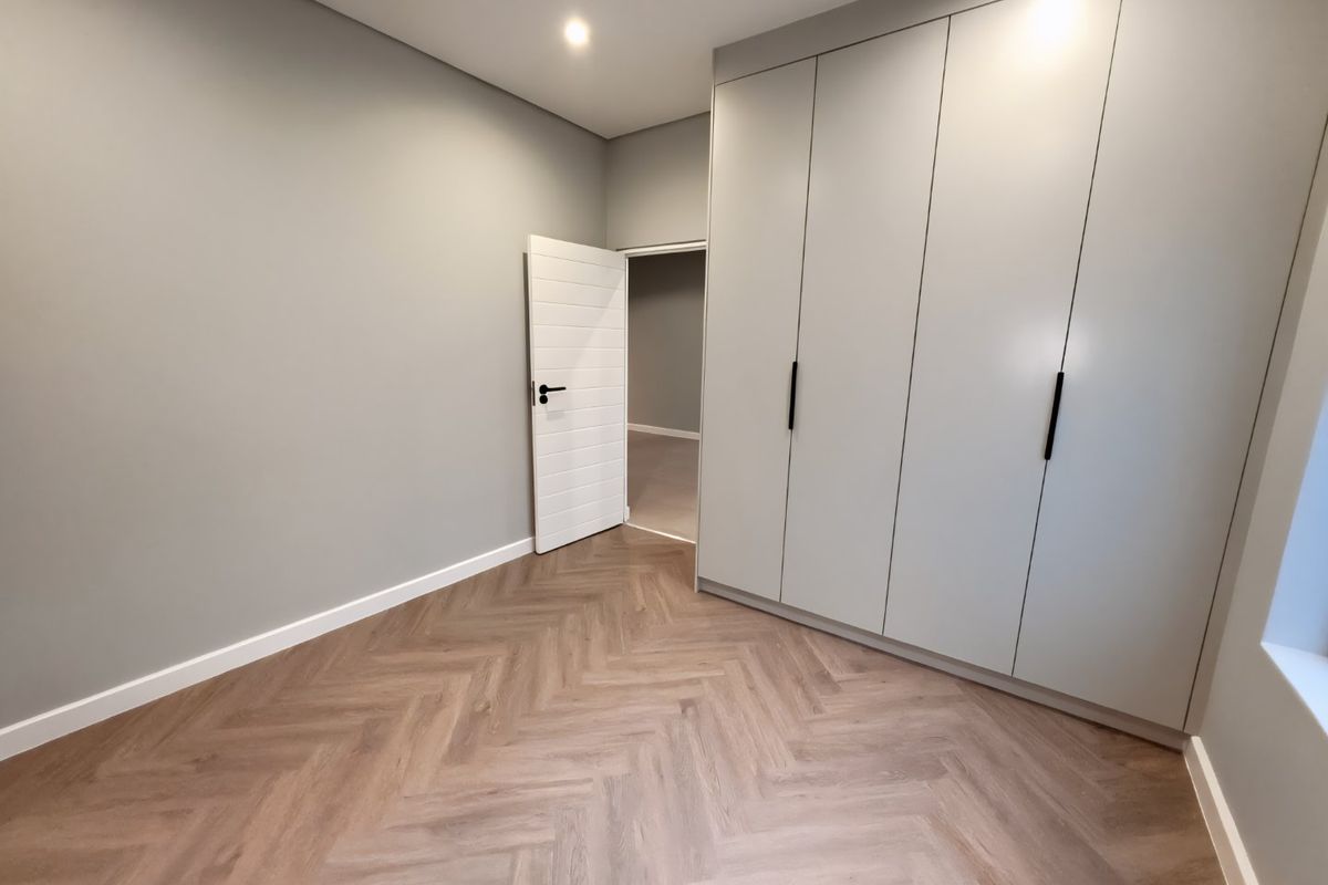 A modern, empty bedroom featuring light grey walls, built-in cupboards, and high-quality herringbone vinyl flooring.