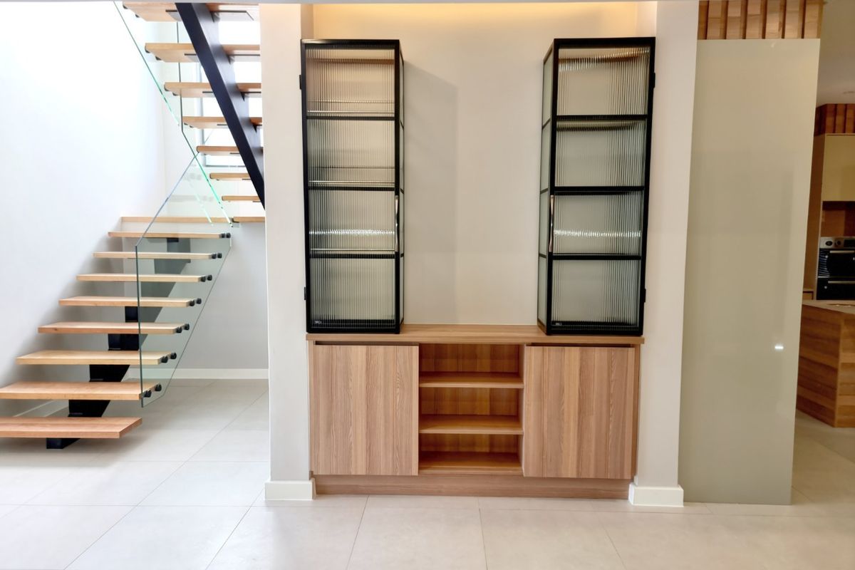 Modern living area featuring a striking floating staircase with glass balustrade and custom built-in timber cabinetry with fluted glass display units.