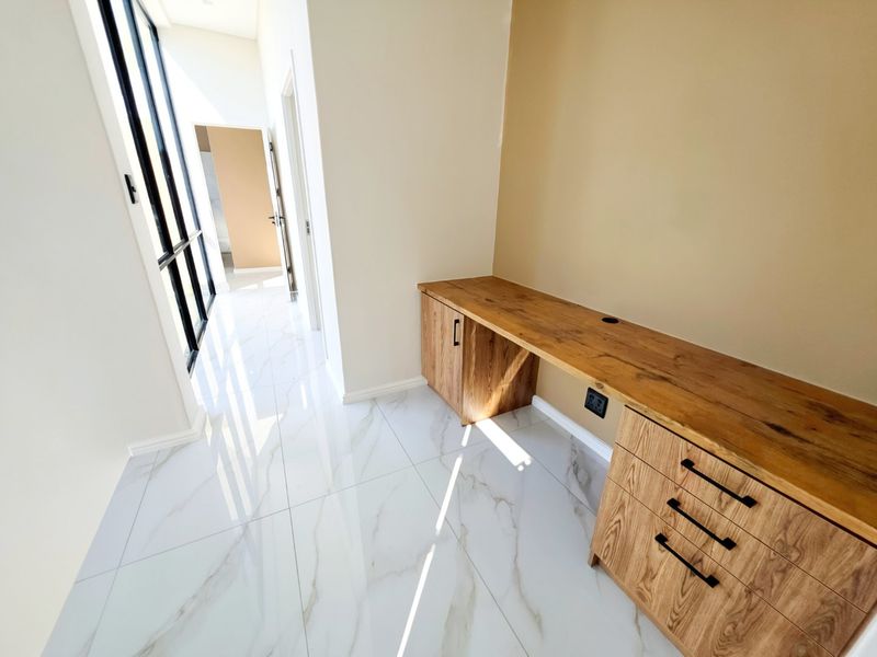 A bright, modern home office space featuring a custom built-in wooden desk with integrated cabinetry and polished marble-look floor tiles.