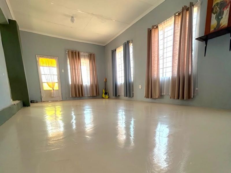 A spacious, empty living room featuring light-colored painted concrete flooring, large windows with security bars, and neutral wall tones.