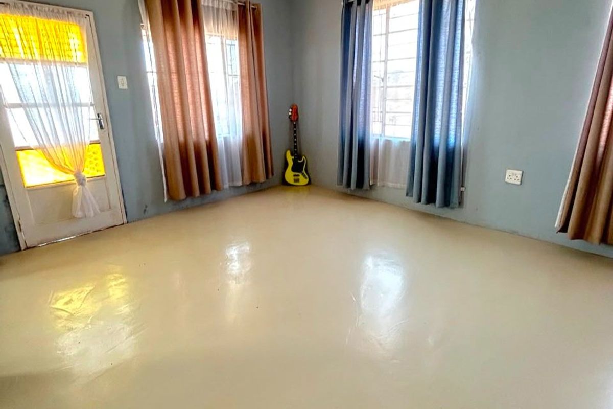 A spacious, empty living room featuring painted concrete floors, large windows with security bars, and a glass panel door leading outside.