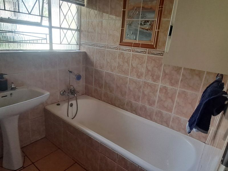 A dated bathroom featuring a pedestal basin, bathtub with mixer tap, and tiled finishes throughout, illuminated by a large leaded glass window.