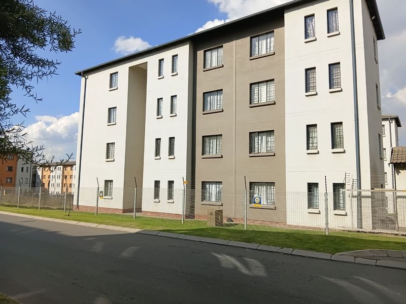 A multi-story apartment building exterior featuring a modern facade with security fencing and window bars, situated along a paved street.