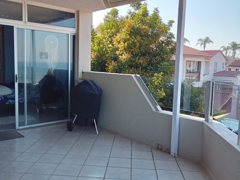 A spacious covered balcony featuring tiled flooring, a glass balustrade, and direct access to the interior through large sliding glass doors.