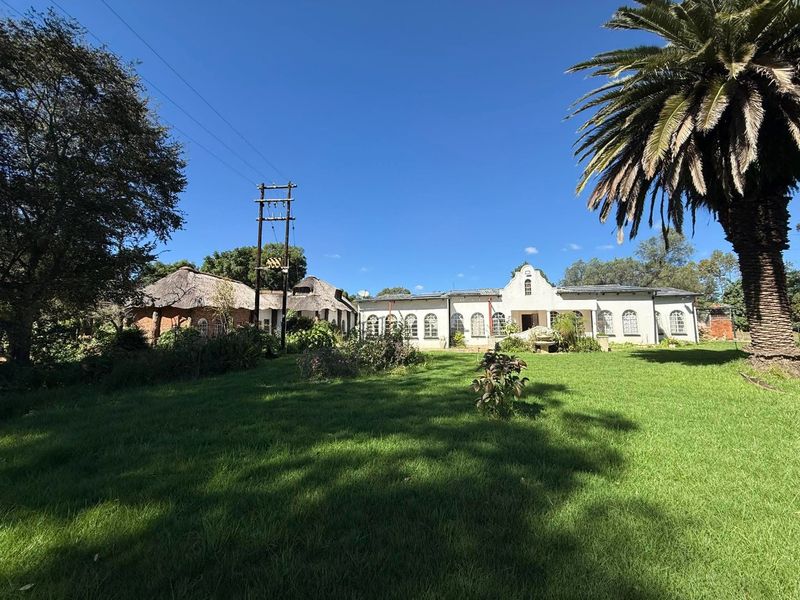 A wide-angle view of a farm property featuring a main residence with arched windows and adjacent thatched-roof outbuildings set within a large garden.